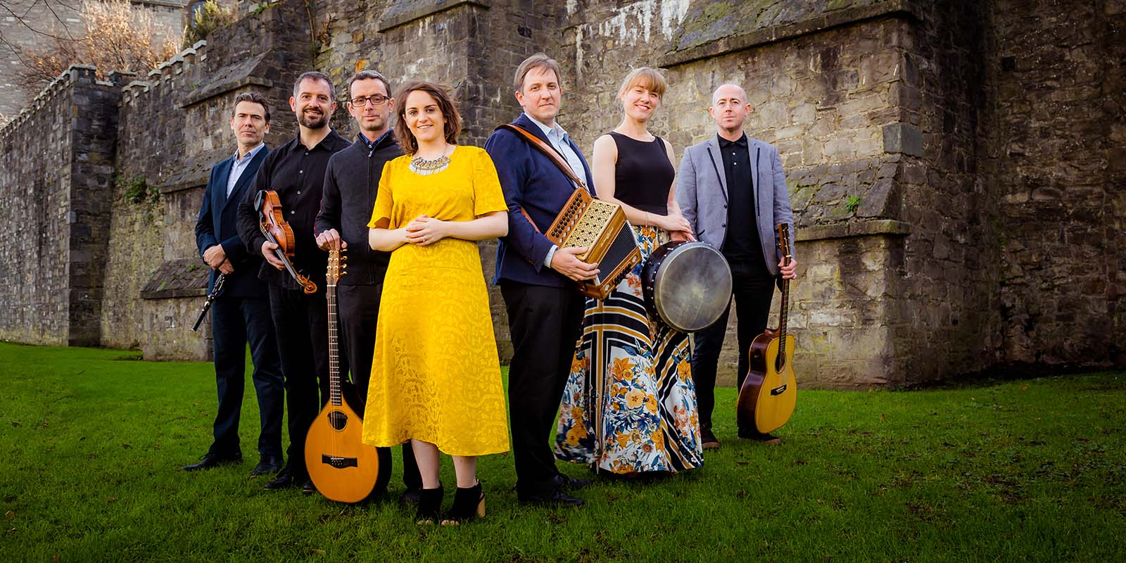 Danú posing with their instruments on green grass with a stone building behind them.