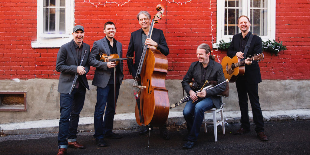 Five men stand and sit in front of a red brick wall, smiling and holding musical instruments including a violin, double bass, flute, uilleann pipes, and guitar. They are dressed in jackets and jeans.