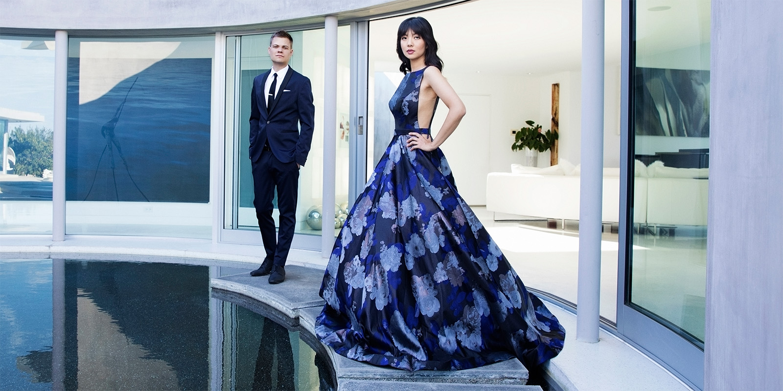 A man in a blue suite and a woman in a flowery blue gown stand beside a swimming pool.