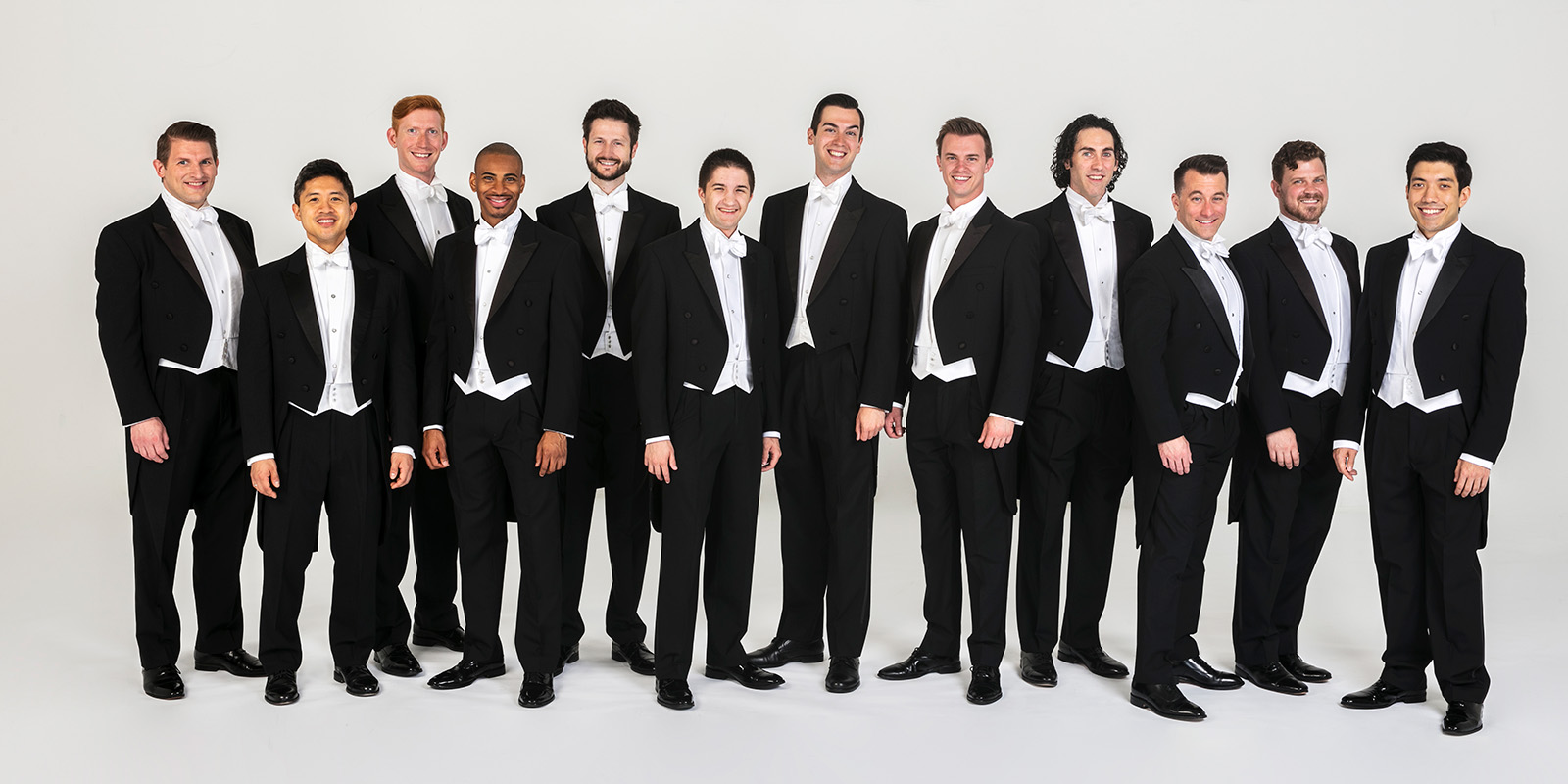 Twelve men in black tuxedos stand and sit in a white studio