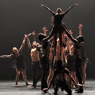 Dancers with outstretched arms and hands lift a woman in the air.