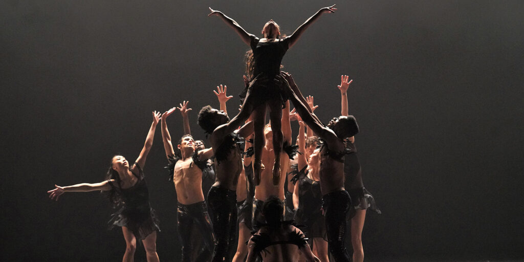 Dancers with outstretched arms and hands lift a woman in the air.