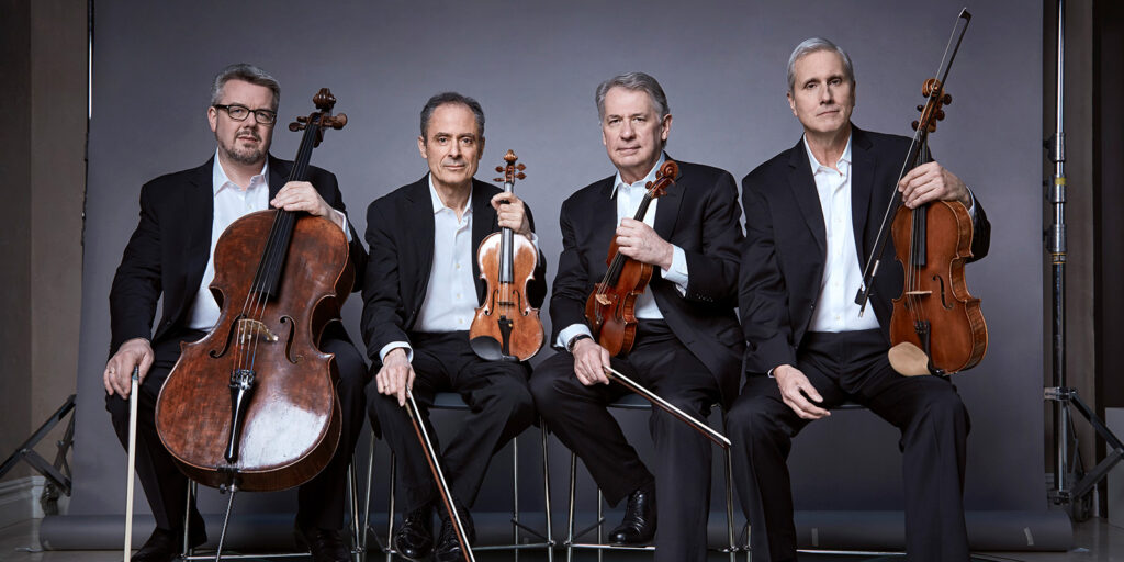 Four men sit in front of a gray backdrop holding string instruments