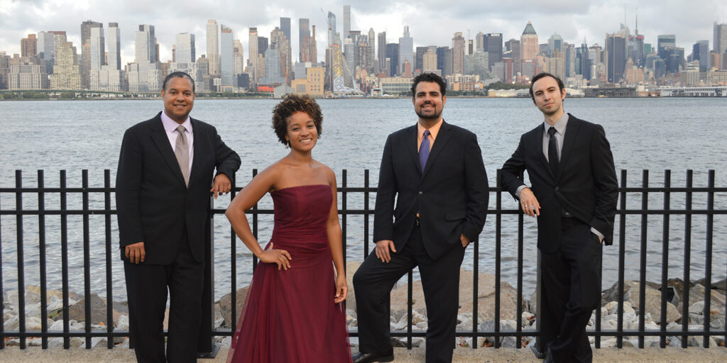 Three men in suits and a woman in a red dress stand in front of a fence across the harbor from New York City.
