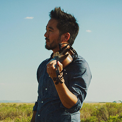 Kishi Bashi stands in a green field holding a violin on his shoulder