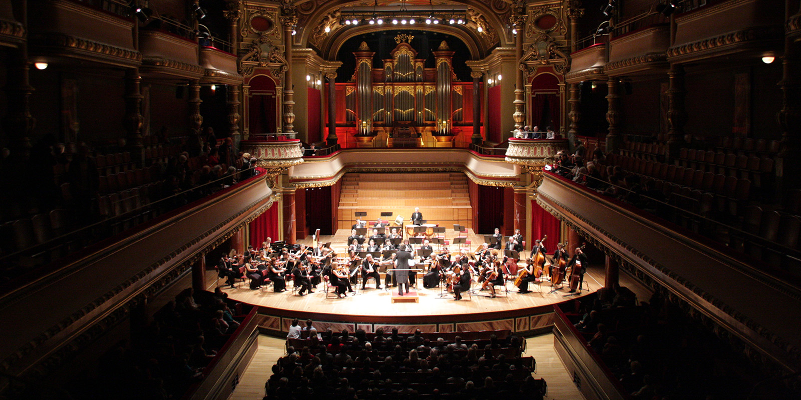 An orchestra performs on stage in an ornate concert hall