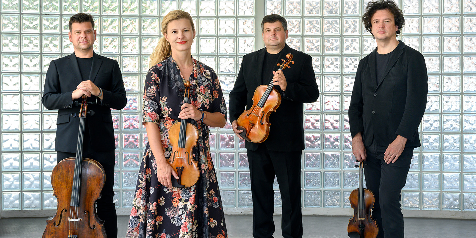 Four musicians hold string instruments while standing in front of a glass wall