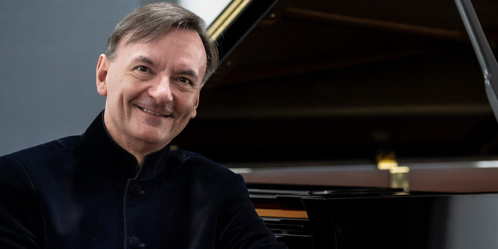 Stephen Hough wears a black suit and sits at a piano