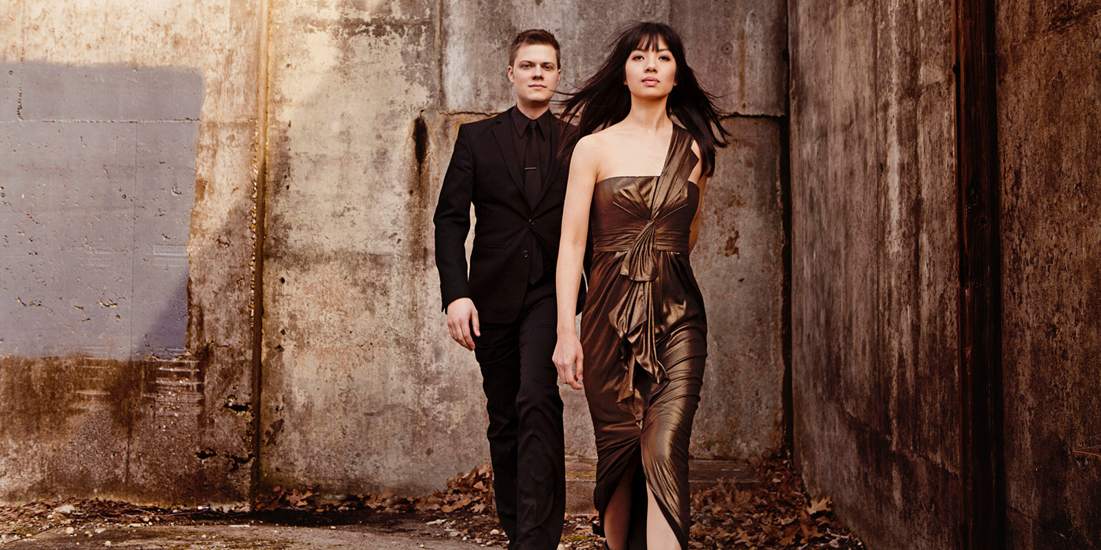 A man in a dark suit and a woman in a brown dress walk in an alleyway