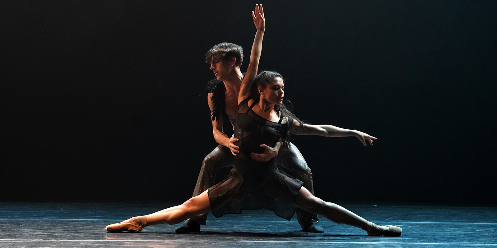 Two dancers in black costumes on on a dark stage.