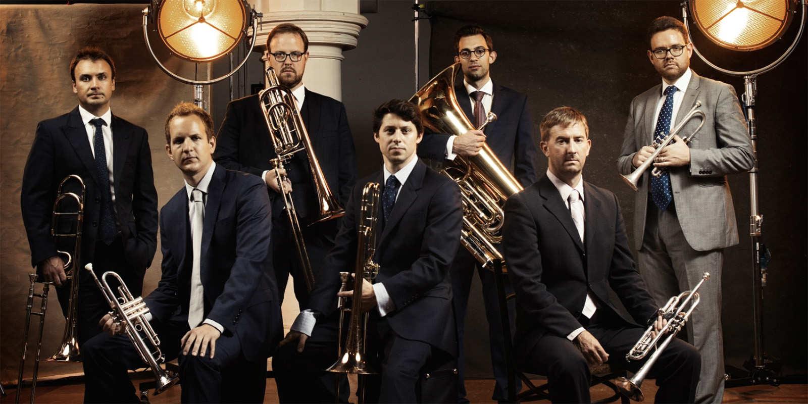 Seven men in suits sit and stand holding brass instruments