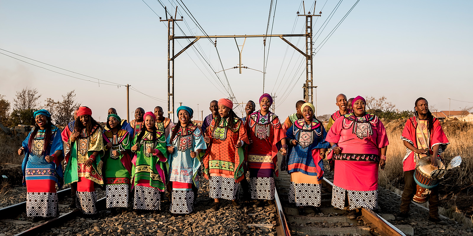 Men and women in colorful clothing sing while standing on railroad tracks