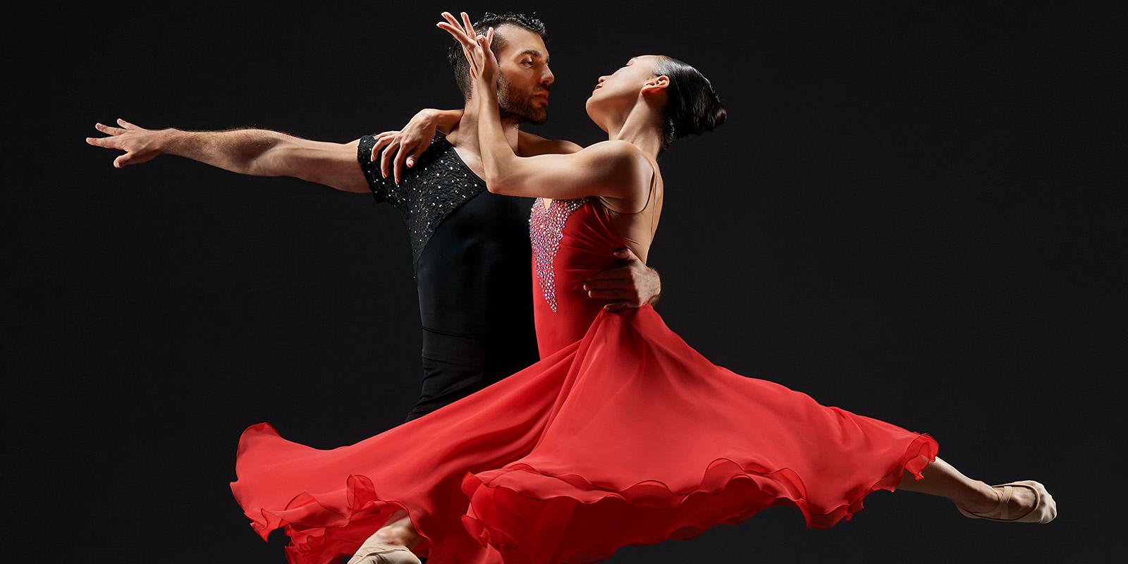 A man in black and a woman in a red dress dance in front of dark background.