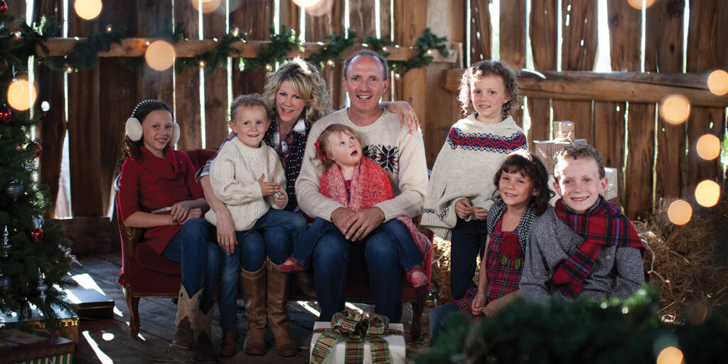 A man and woman and five children wear festive sweaters and sit in a barn near a Christmas tree