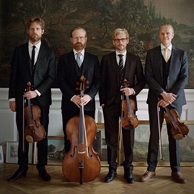 Four men in dark suits stand in a line holding violins, a viola, and a cello