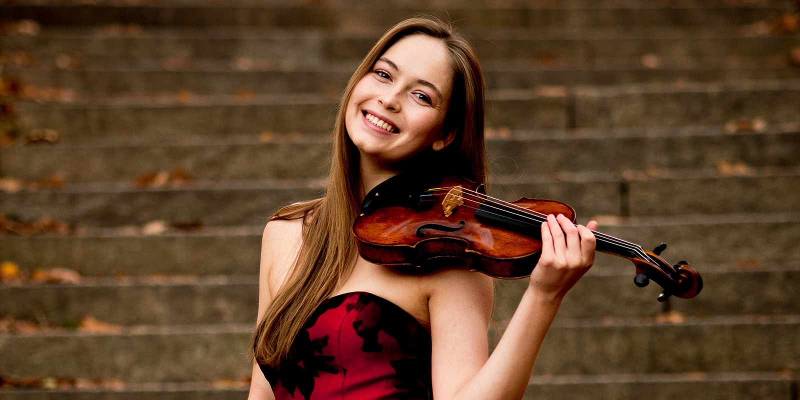 A woman with long brown hair wears a red and black dress and holds a violin on her shoulder.