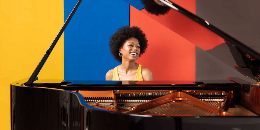 A woman sits at a black grand piano in front of a colorful striped wall.