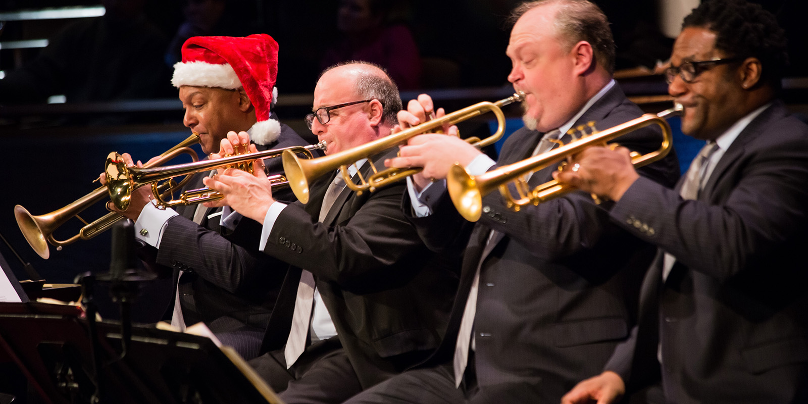 Two men play trumpets while looking at music. One wears a red Santa Claus hat.