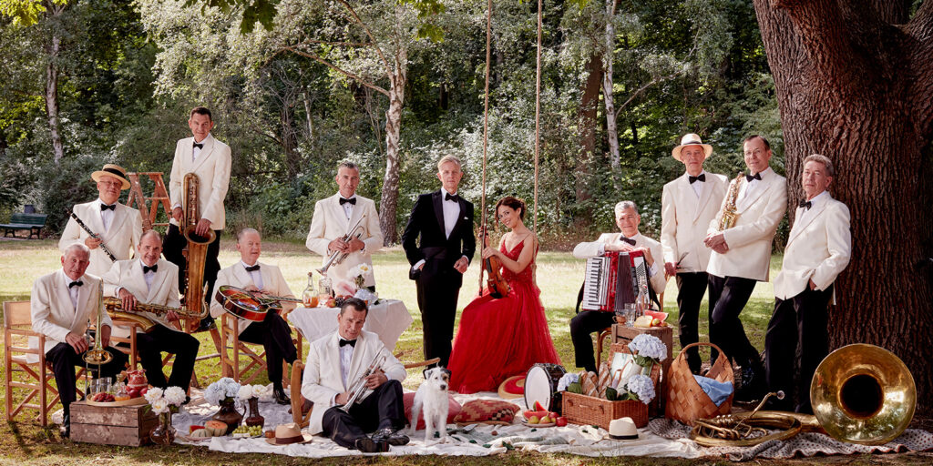 A group of musicians hold instruments and sit on a lawn in front of a large tree.