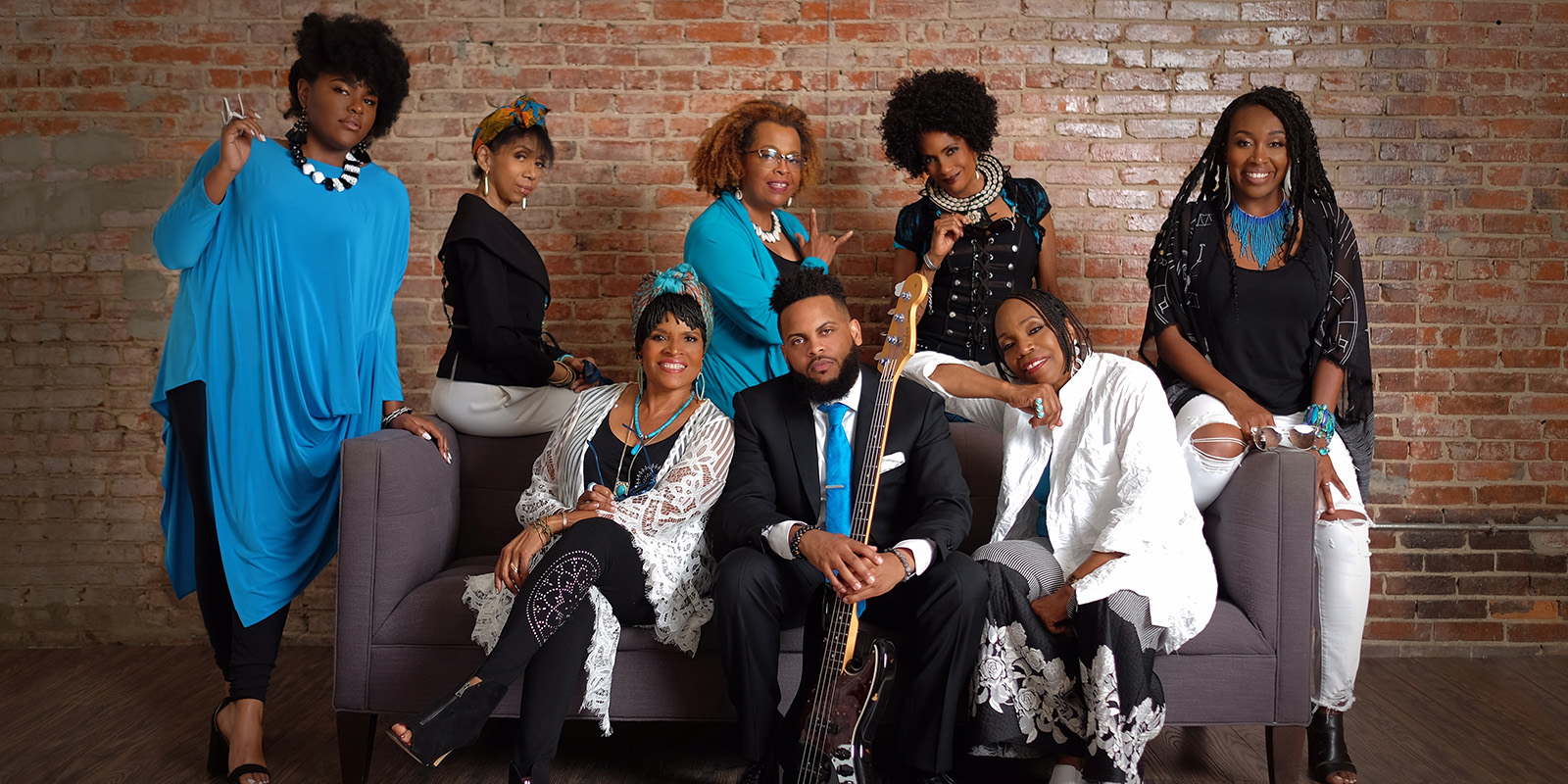 Seven women and one man in black and blue clothing sit on and around a gray couch in front of a red brick wall.