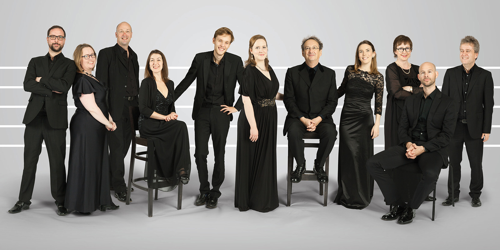 Eleven men and women wearing black suites and dresses stand and sit on tall stools in front of a white and grey background.