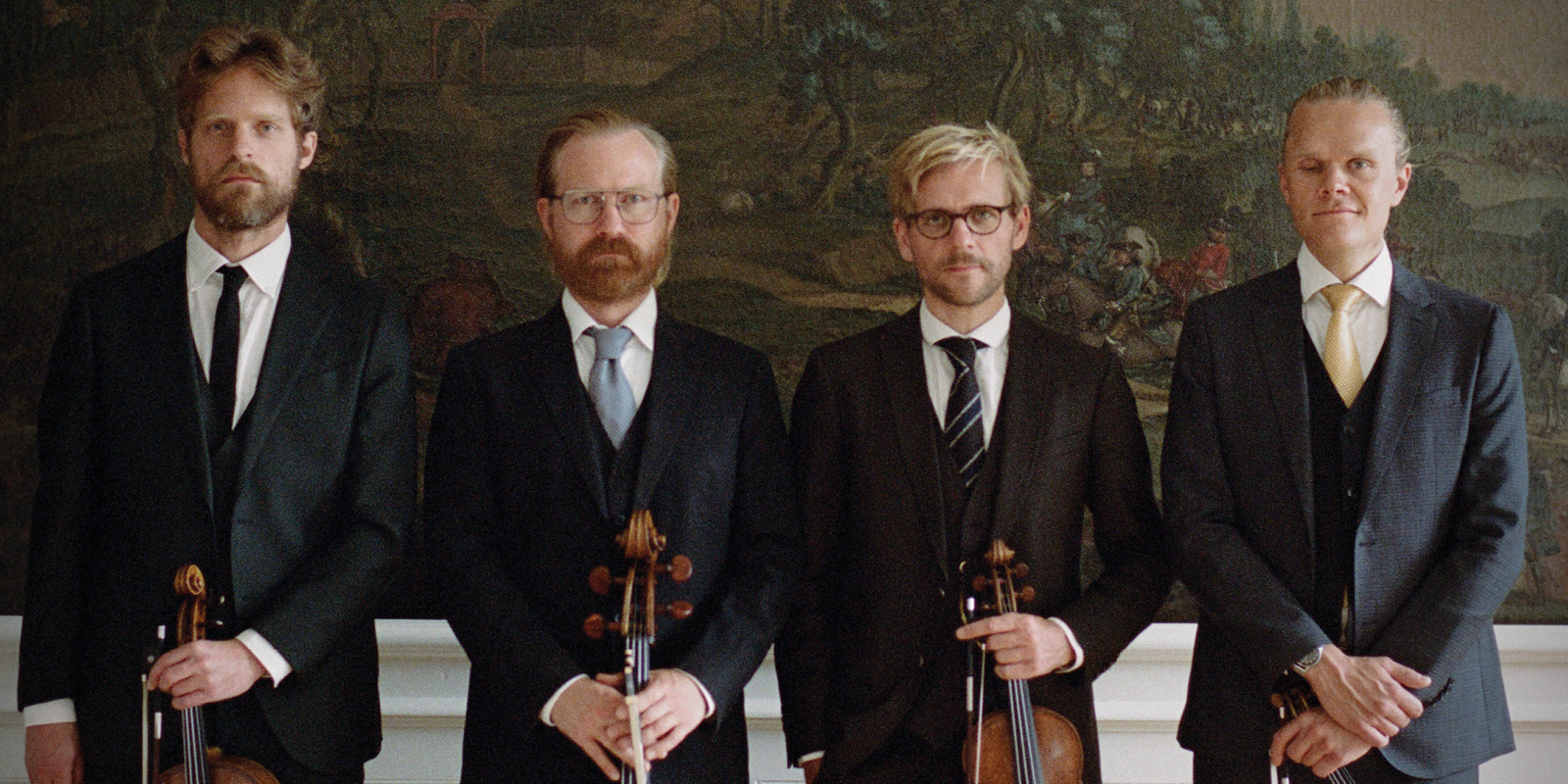 Four men in dark suits stand shoulder to should holding string instruments in front of a large mural.