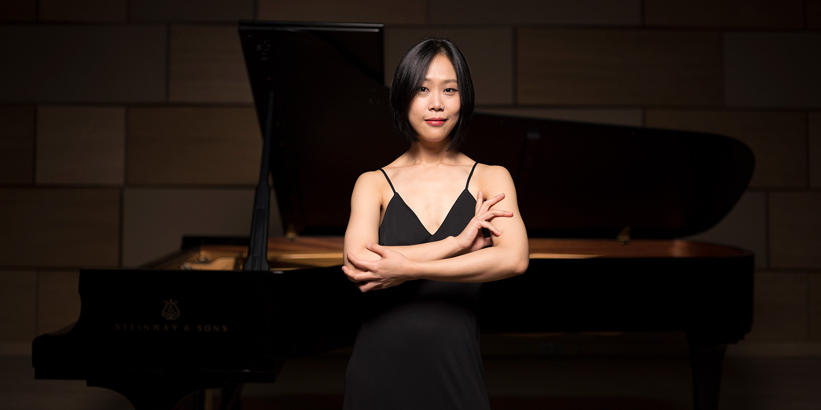 A woman with short dark hair stands in front of a black grand piano with her arms crossed.