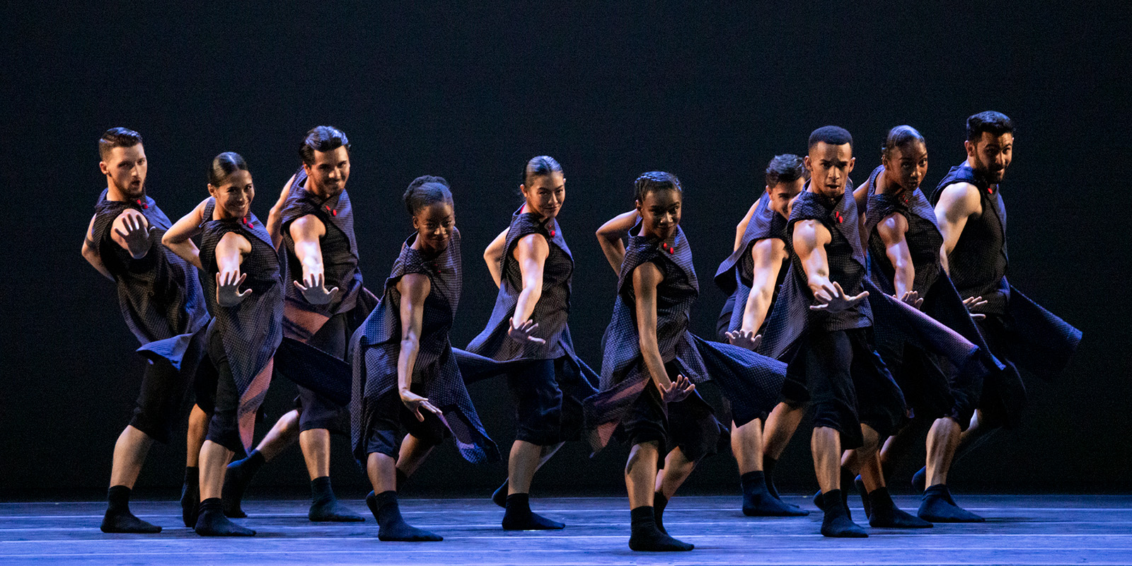 Dancers in dark clothing dance in a line on a dark stage.