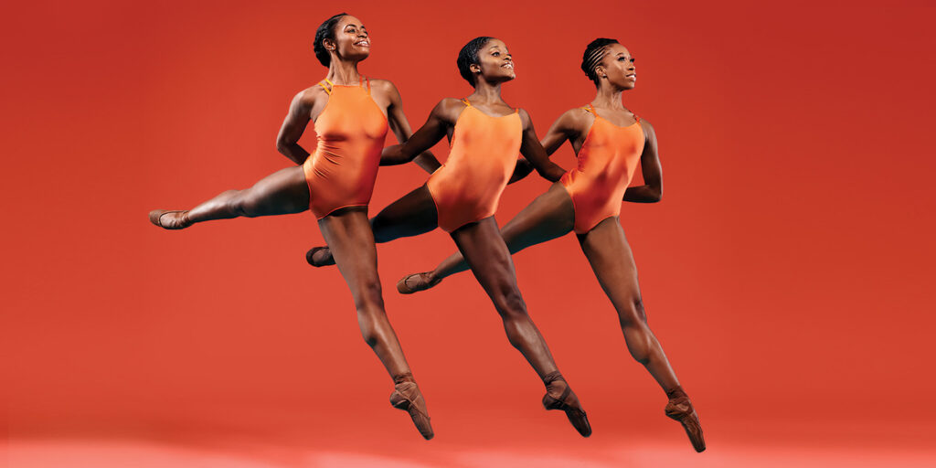 Three women in orange leotards leap in the air.