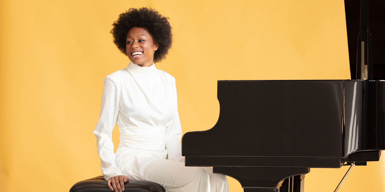 A woman in a white suit sits at a black piano in front of a bright yellow wall.
