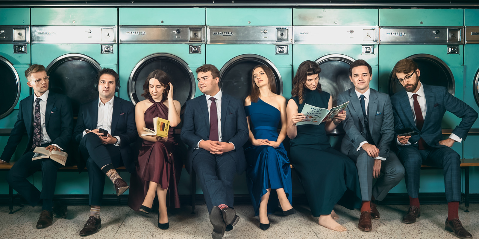 Eight men and women in formal attire sit in a laundromat.