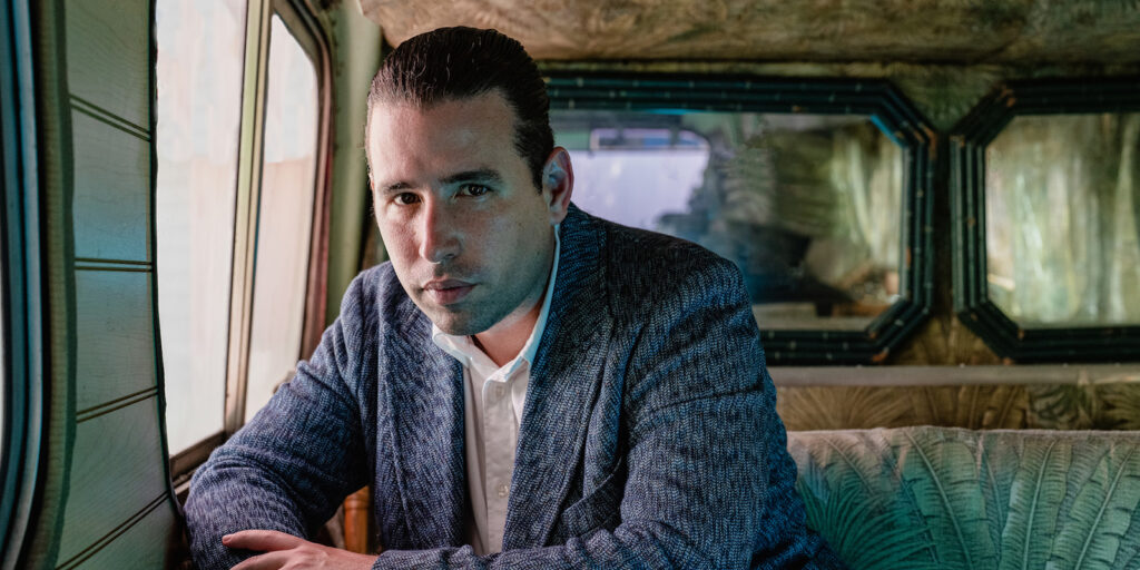 A man in a vintage bus leans forward and looks at the camera. He wears a light blue jacket.