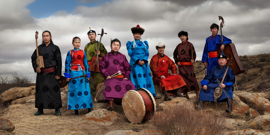 Men and women in traditional Mongolian clothing sit on a rocky, arid hillside holding traditional instruments.