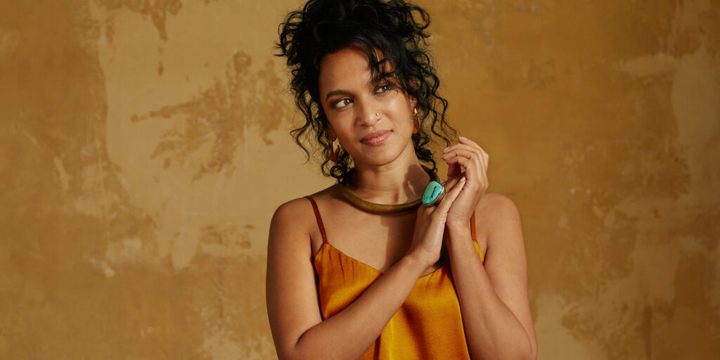 A woman wears a golden sleeveless top and wears a large turquoise ring. She stands in front of a textured golden wall.