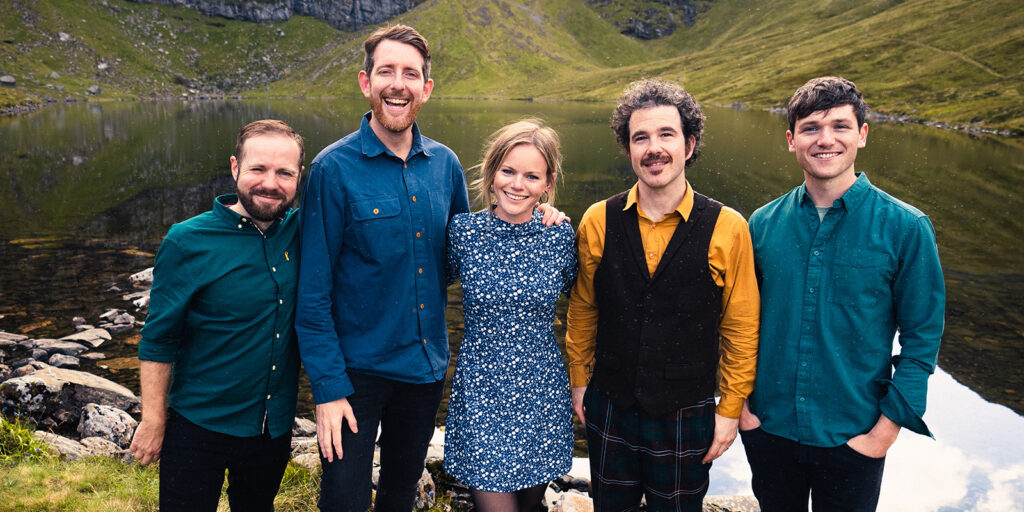 Four men and one woman in colorful shirts and dark pants stand on a rugged, green hill in the highlands of Scotland.