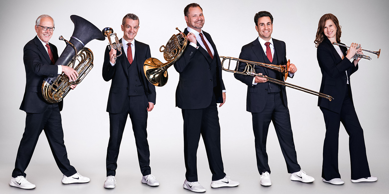 Four men and a woman in dark suits and white sneakers stand in a line holding brass instruments.
