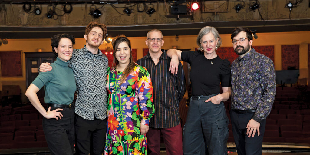 A group of musicians stand close to each other on a bare stage.