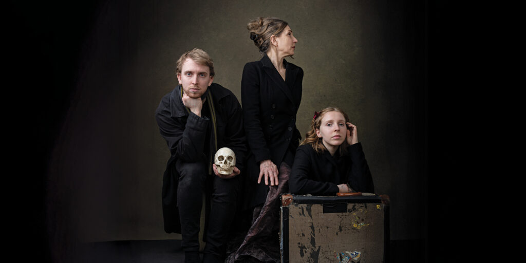 A man and two women sit and gaze forward or off to the side. The young woman leans on an old brown suitcase. They are wearing dark clothing. The women in the center holds a human skull in her hands.