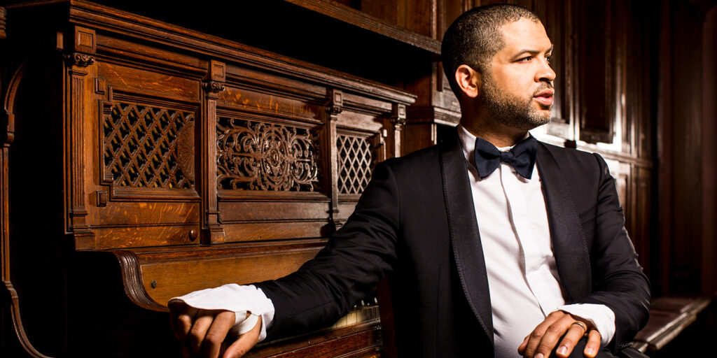 A man in a black suit sits at a vintage piano.