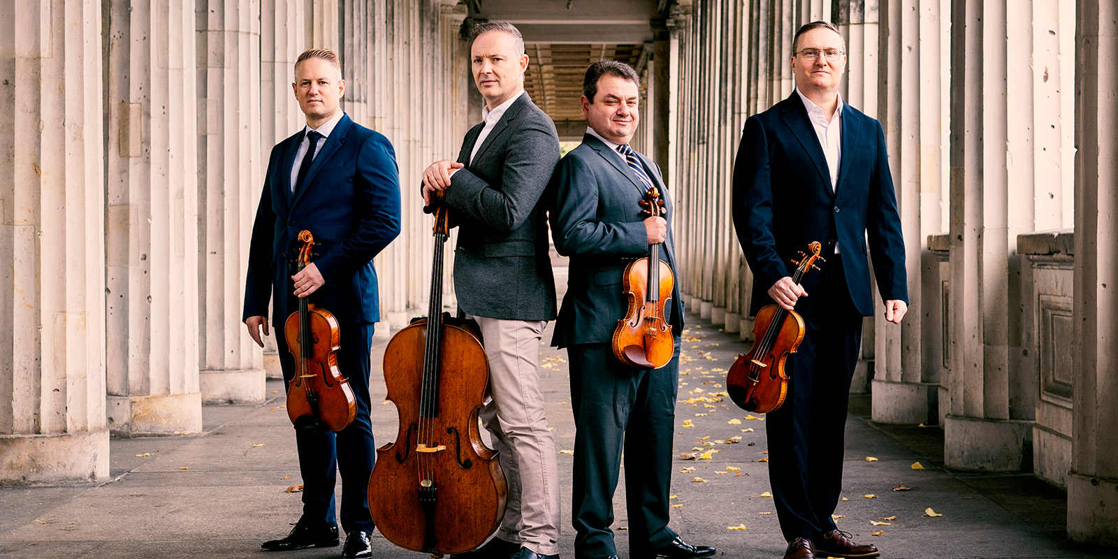 Four men in dark suits stand in an outdoor promenade holding their string instruments.