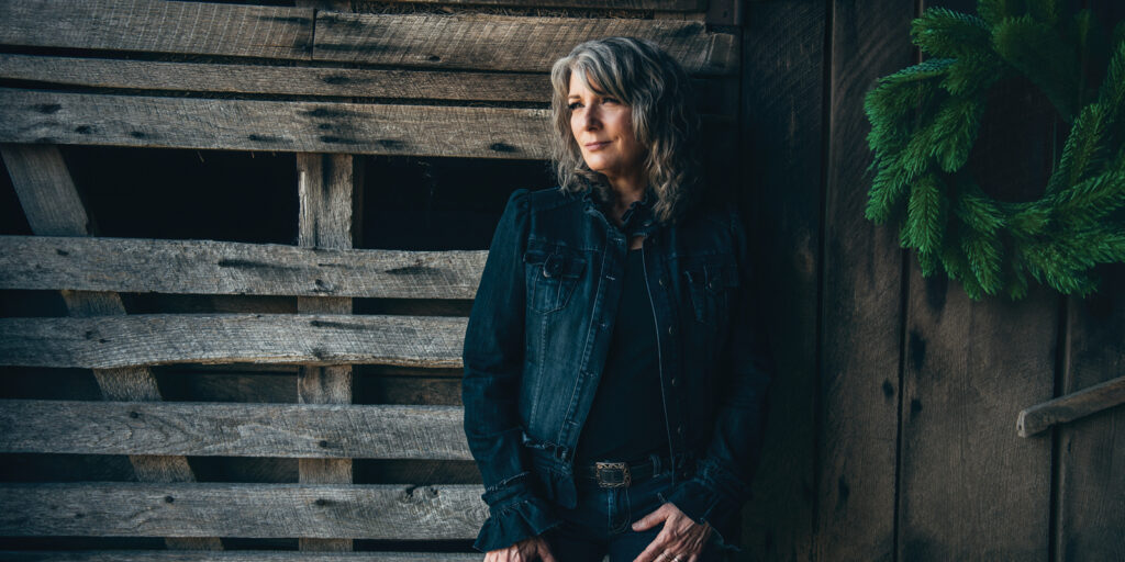 A woman with shoulder length wavy hear leans against a rough wooden fence or barn. She is wearing a dark blue denim jacket.