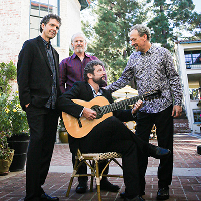 Three men stand around a chair in an outdoor courtyard. One man, holding a guitar, sits on the chair and looks up at the men beside him.