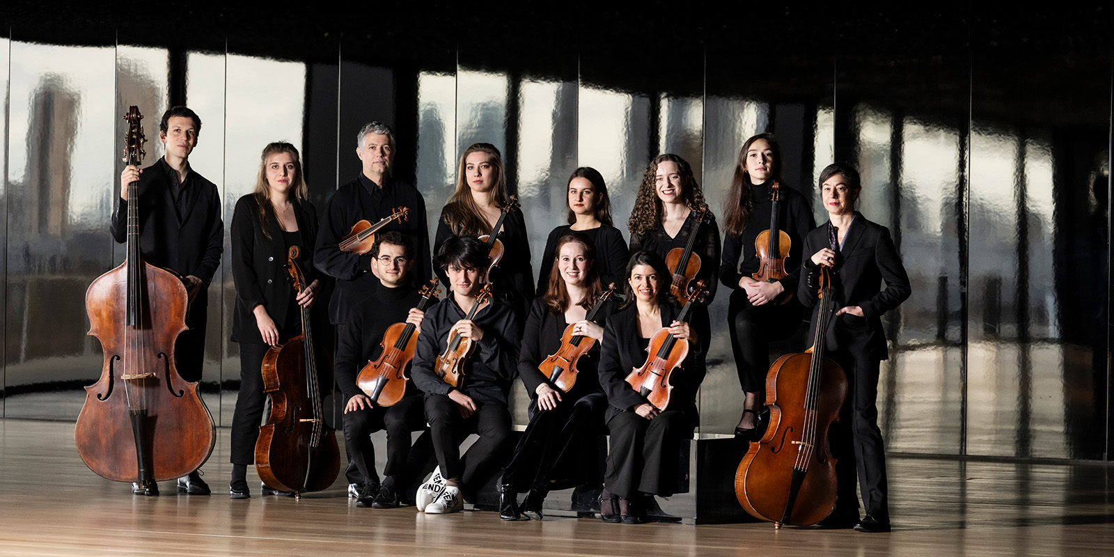Twelve musicians in black clothing sit and stand in a large empty studio in front of windows while holding string instruments.