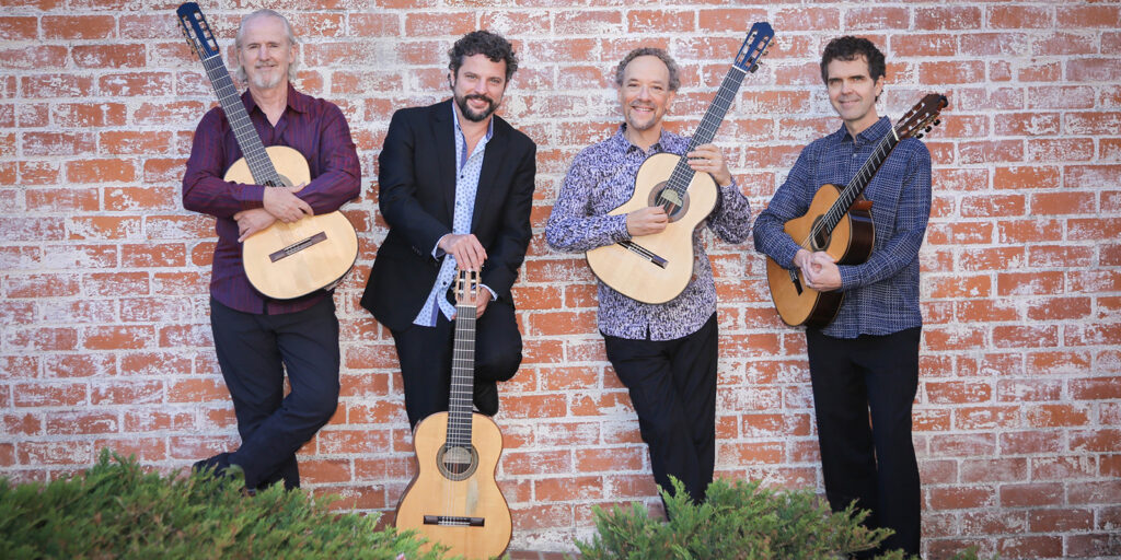 Four men hold guitars and stand in front of a red brick wall.