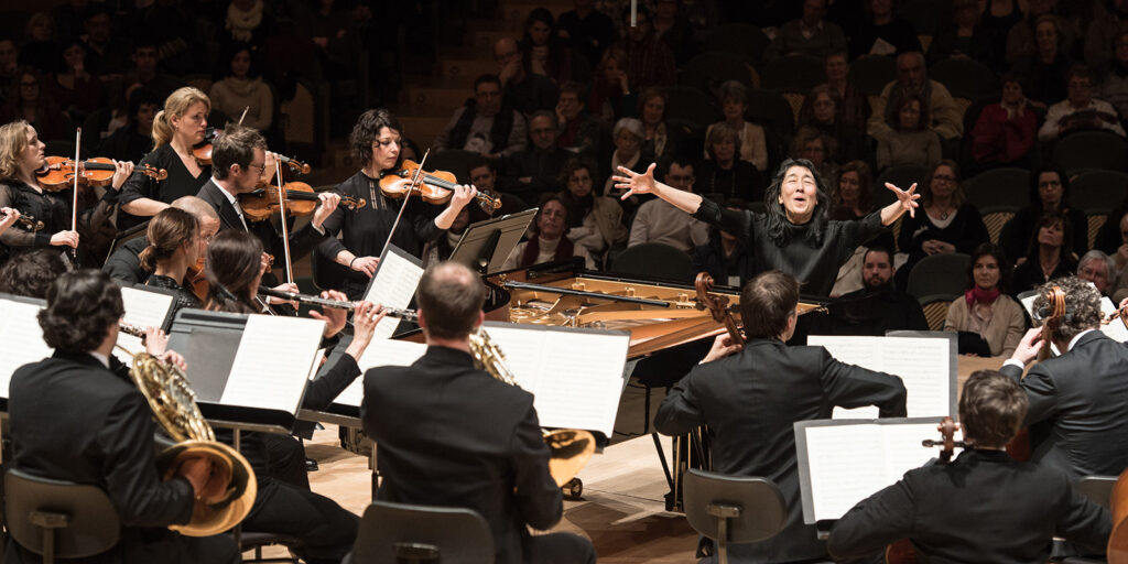 A woman with outstretched arms sits on a stage at a piano with her back to the audience surrounded by musicians playing instruments.