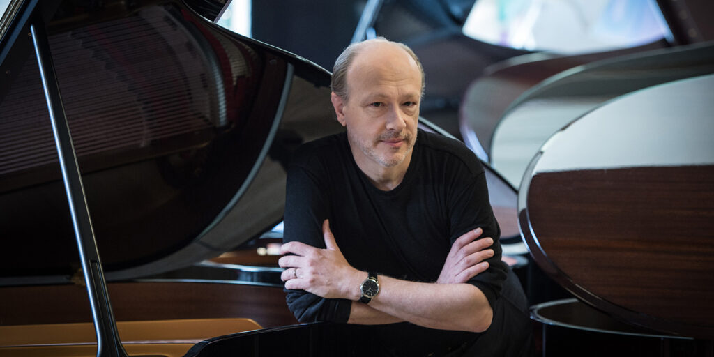 A man in a white shirt leans forward and rests his arms on a black piano, surrounded by more pianos.