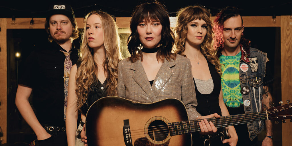 A woman holds a guitar. She is surrounded by two men and two women. They all wear eclectic clothing.