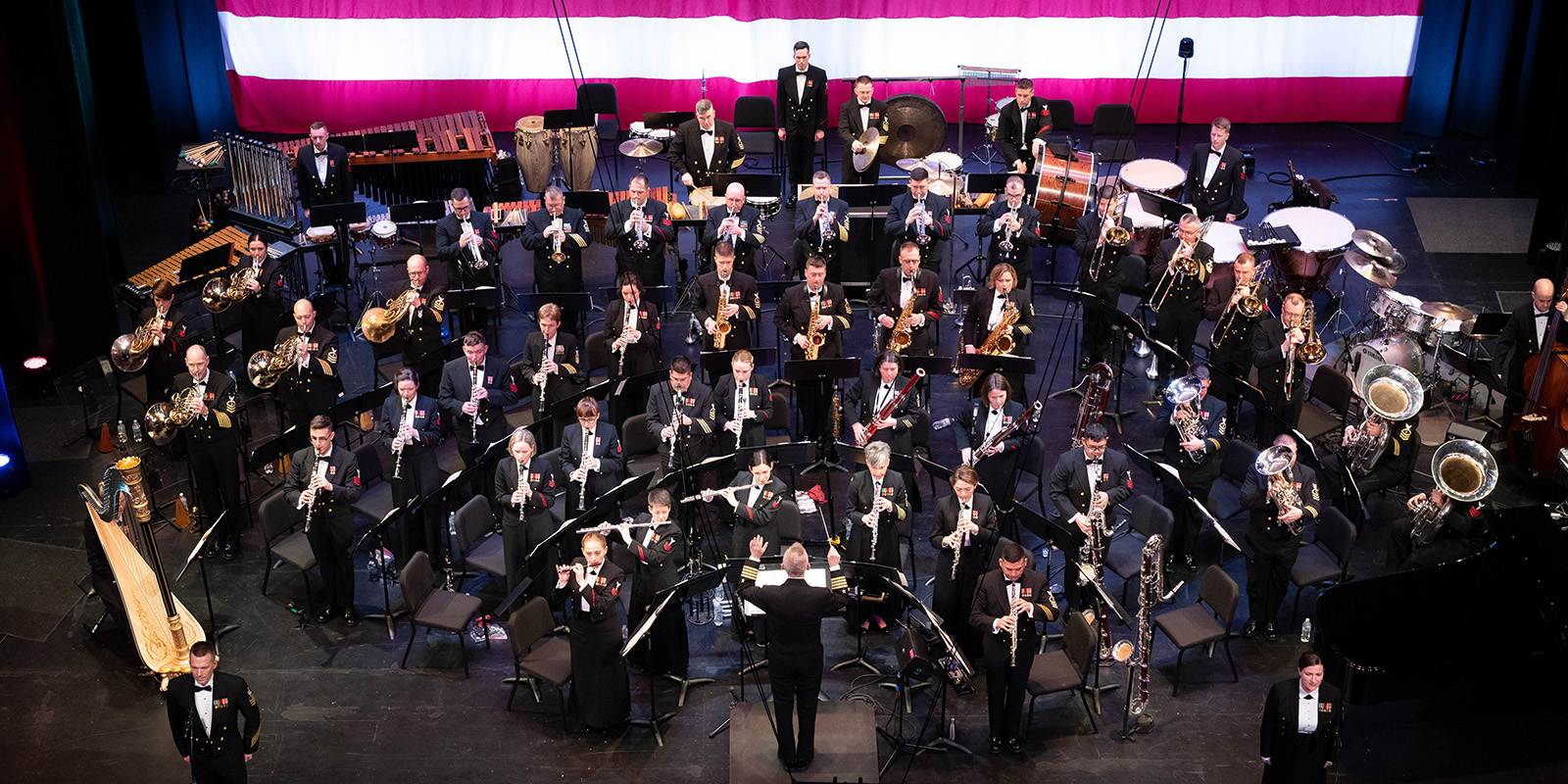 A military band performs on a dark stage in front of a large U.S. flag.