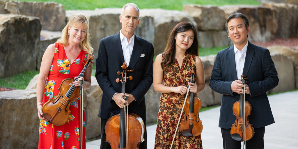 Four musicians hold string instruments while standing in a garden filled with large stones and green grass