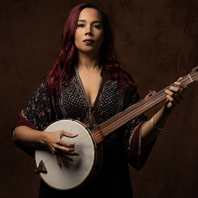 A woman with straight reddish hair faces the camera and holds a banjo in her arms.